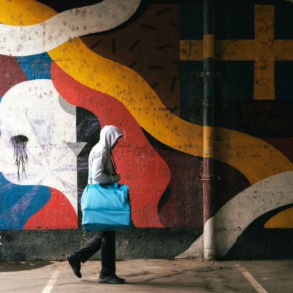 Homme en sweat à capuche marchant avec un sac bleu devant un mur coloré illustrant les bienfaits de la boxe française