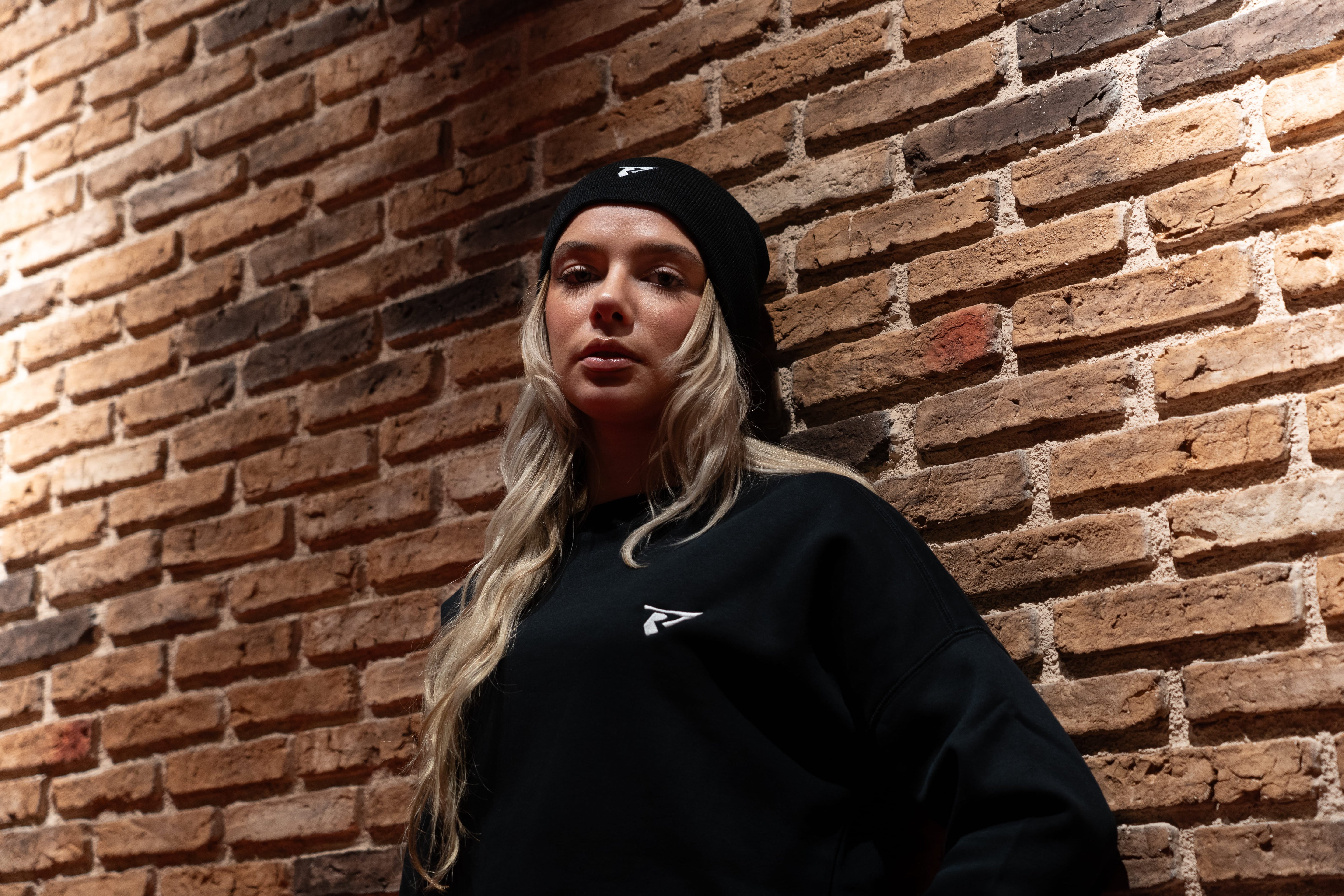 Femme portant un sweat noir de la collection EVO T-shirts et Sweats sport femme avec un bonnet assorti devant un mur en briques